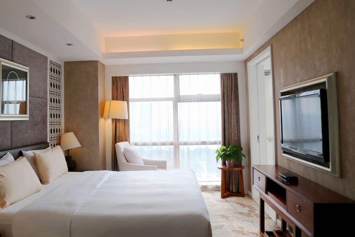 Ein Hotelzimmer mit einem großen Bett, einem Fernseher, einem Schreibtisch mit Lampe, einem Sessel und Blick ins Freie durch das Fenster.