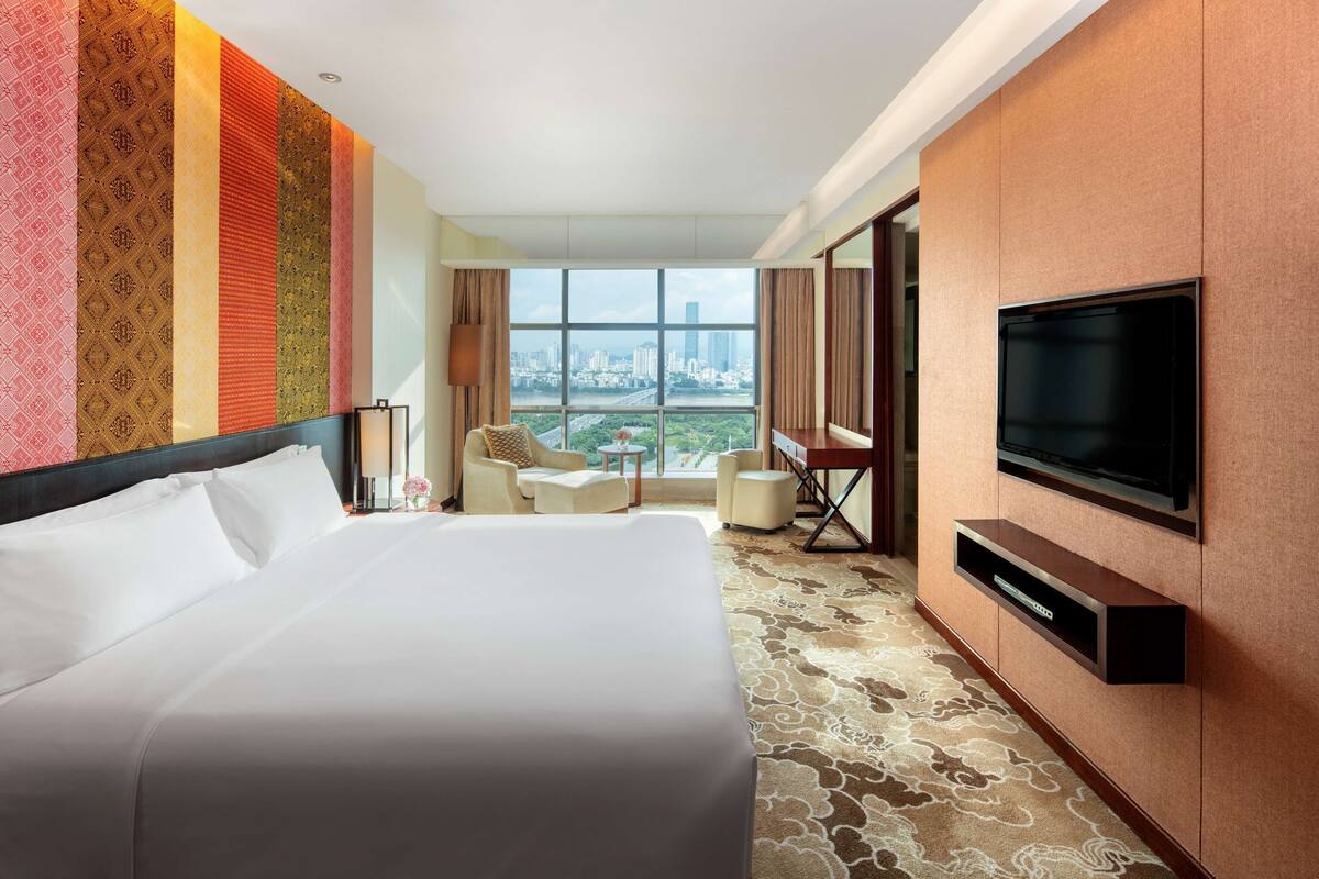 Ein Hotelzimmer mit einem großen Bett, einem an der Wand montierten Fernseher, einem Schreibtisch und einem Sessel.