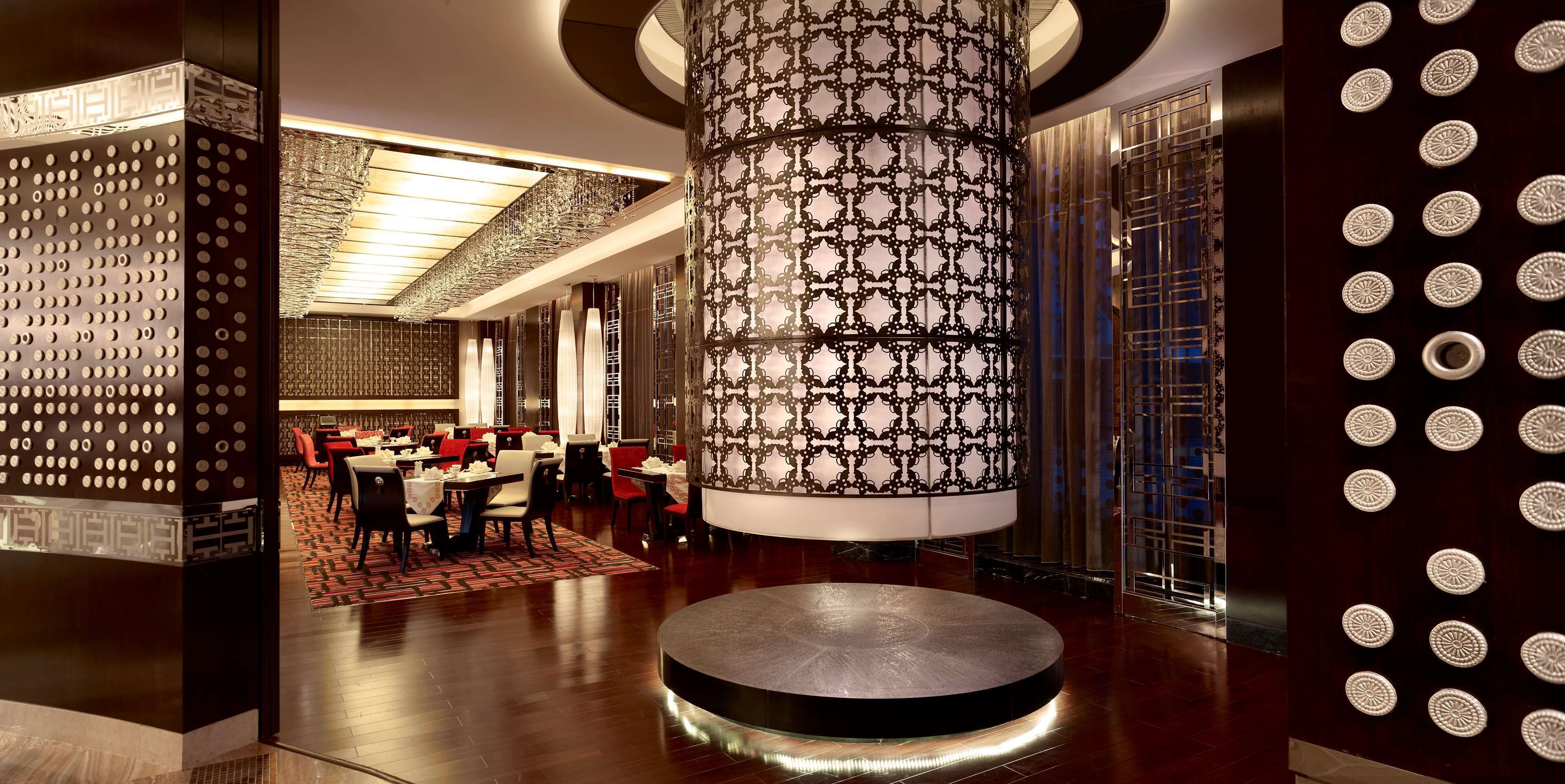 A modern hotel lobby with a central circular light fixture, patterned walls, and a seating area with tables and chairs.