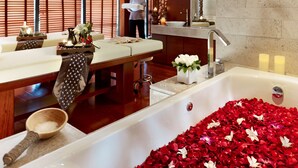 Um quarto de spa com uma banheira grande cheia de pétalas de rosa, uma mesa de massagem e uma área de recepção com alguém atendendo.