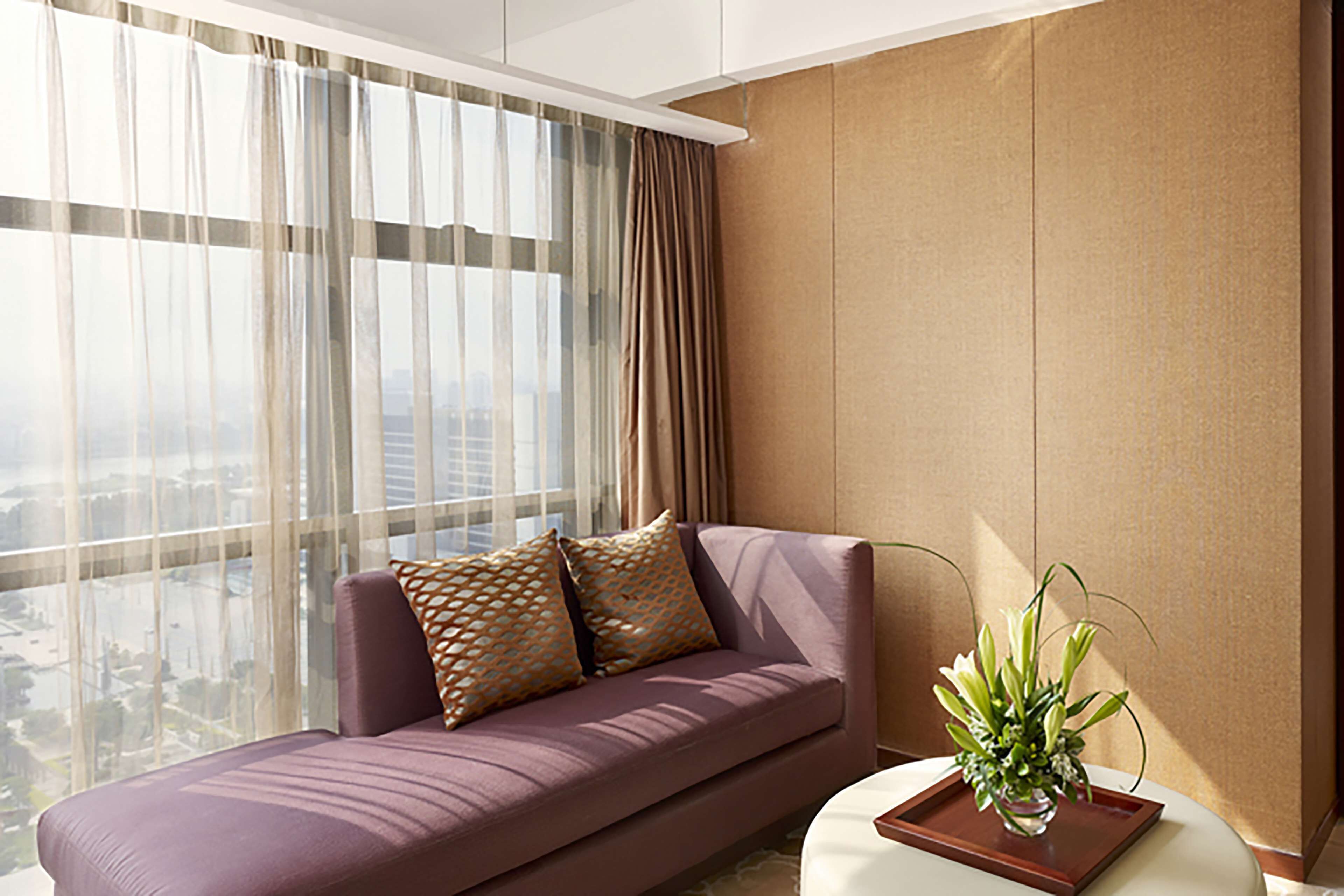 A modern living room with a purple sofa, a vase with flowers, and a city view through sheer curtains.