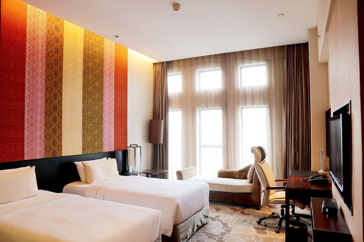 Ein Hotelzimmer mit zwei Betten, einem Schreibtisch mit Stuhl, einem Fernseher und großen Fenstern mit Vorhängen.