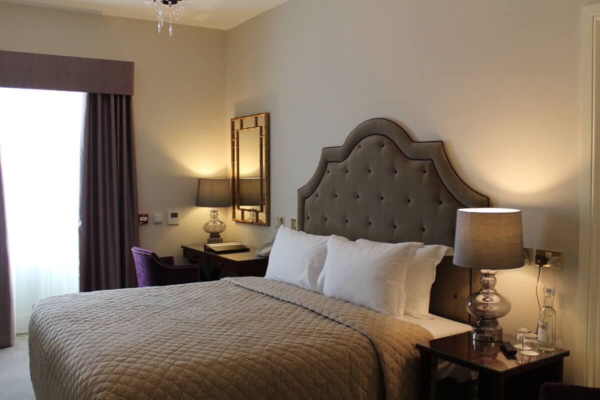 Une chambre d’hôtel avec un grand lit, une table de chevet avec une lampe, une chaise, un miroir et une fenêtre avec des rideaux.