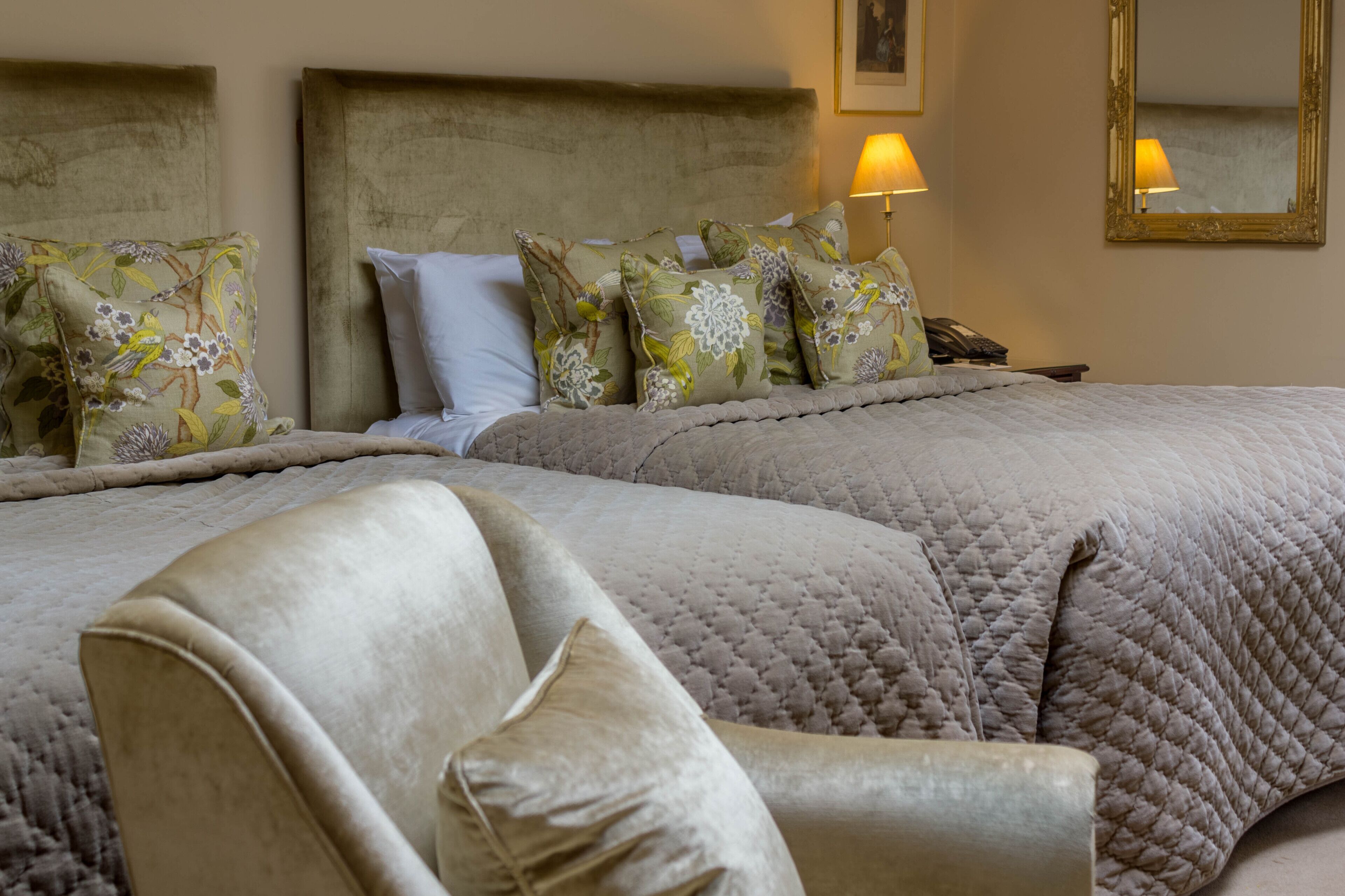 A hotel room with a large bed, multiple pillows, a chair, and a bedside table with a lamp and phone.
