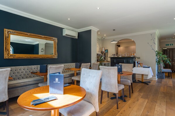 Un vestíbulo de hotel con una pared oscura, un espejo grande, una zona de estar con un sofá y sillas, una mesa de madera con una carpeta y una cocina al fondo.