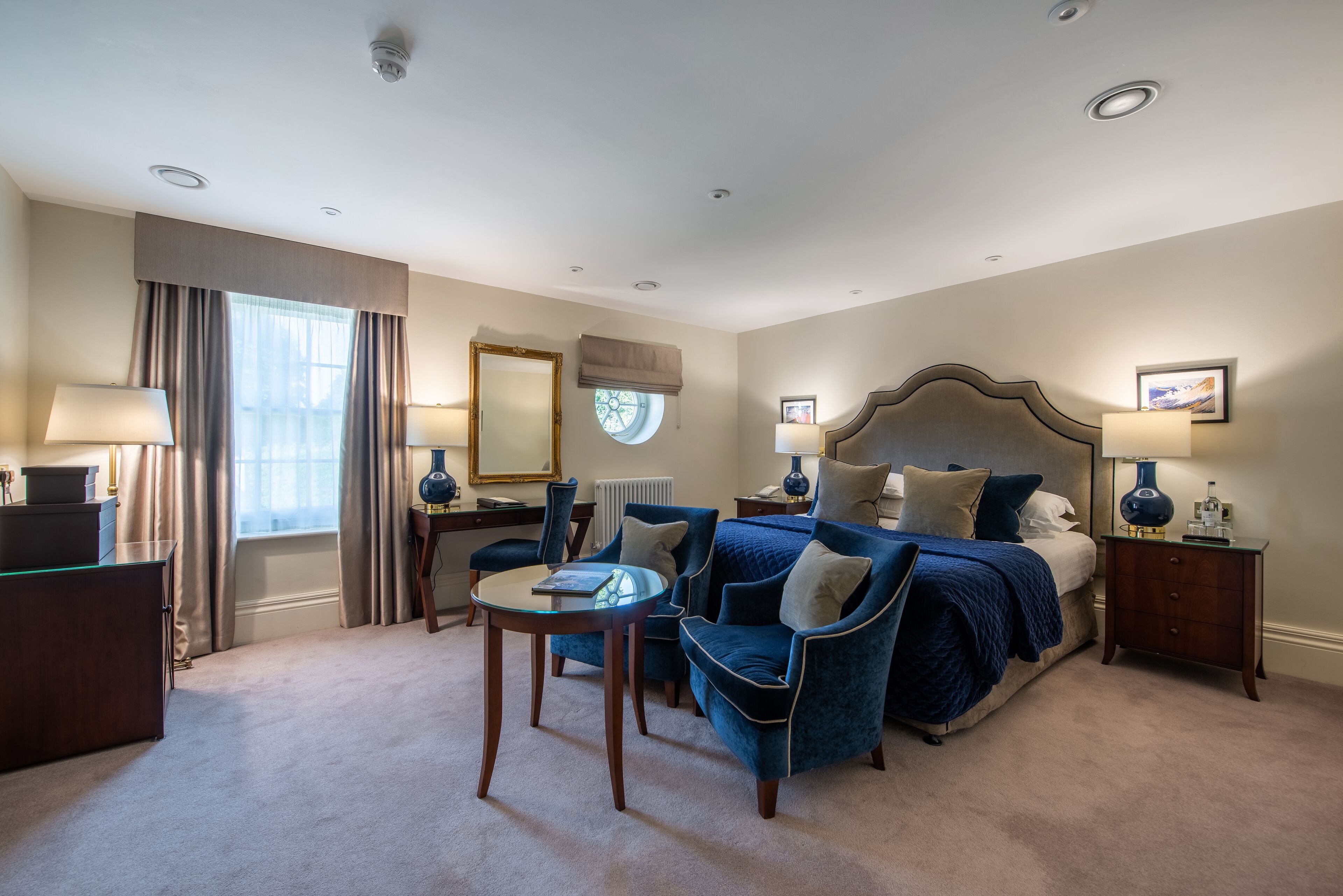 A hotel room with a large bed, two armchairs, a small table, a desk, and a window with curtains.