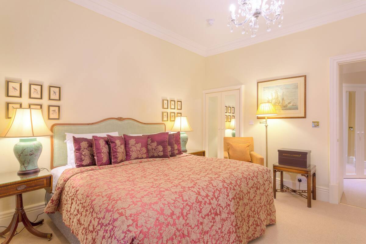 Une chambre à coucher bien rangée, avec un grand lit, deux tables de chevet avec des lampes, une chaise et un tableau encadré au mur.