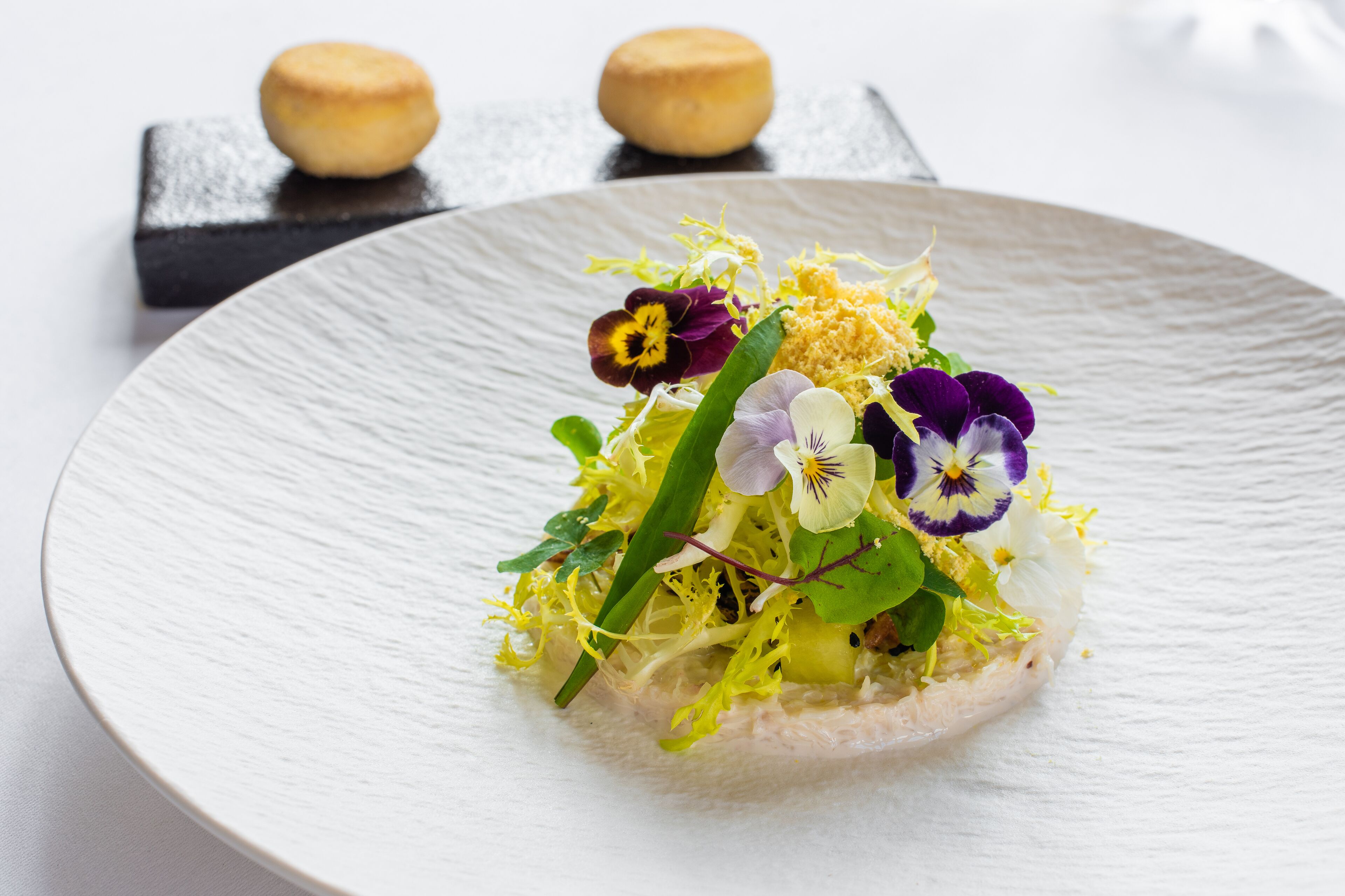 A white plate with a food arrangement featuring greens, flowers, and a small pastry.