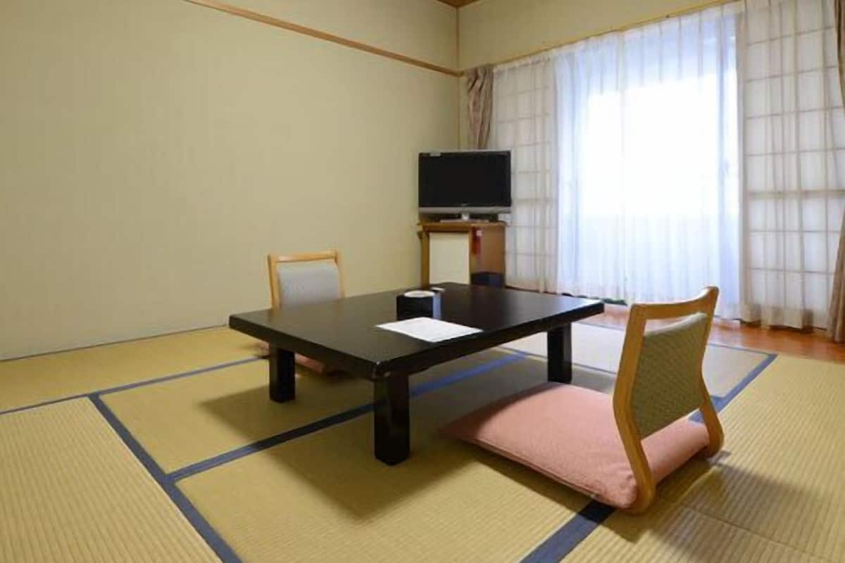 A room with tatami mat flooring, a low table, chairs, a TV, and a window with curtains.