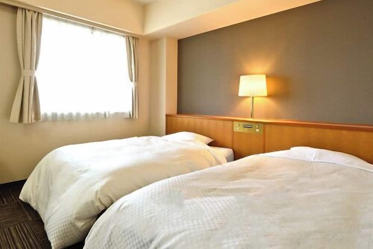 A hotel room with two beds, a window with curtains, a wall-mounted lamp, and a wooden headboard.