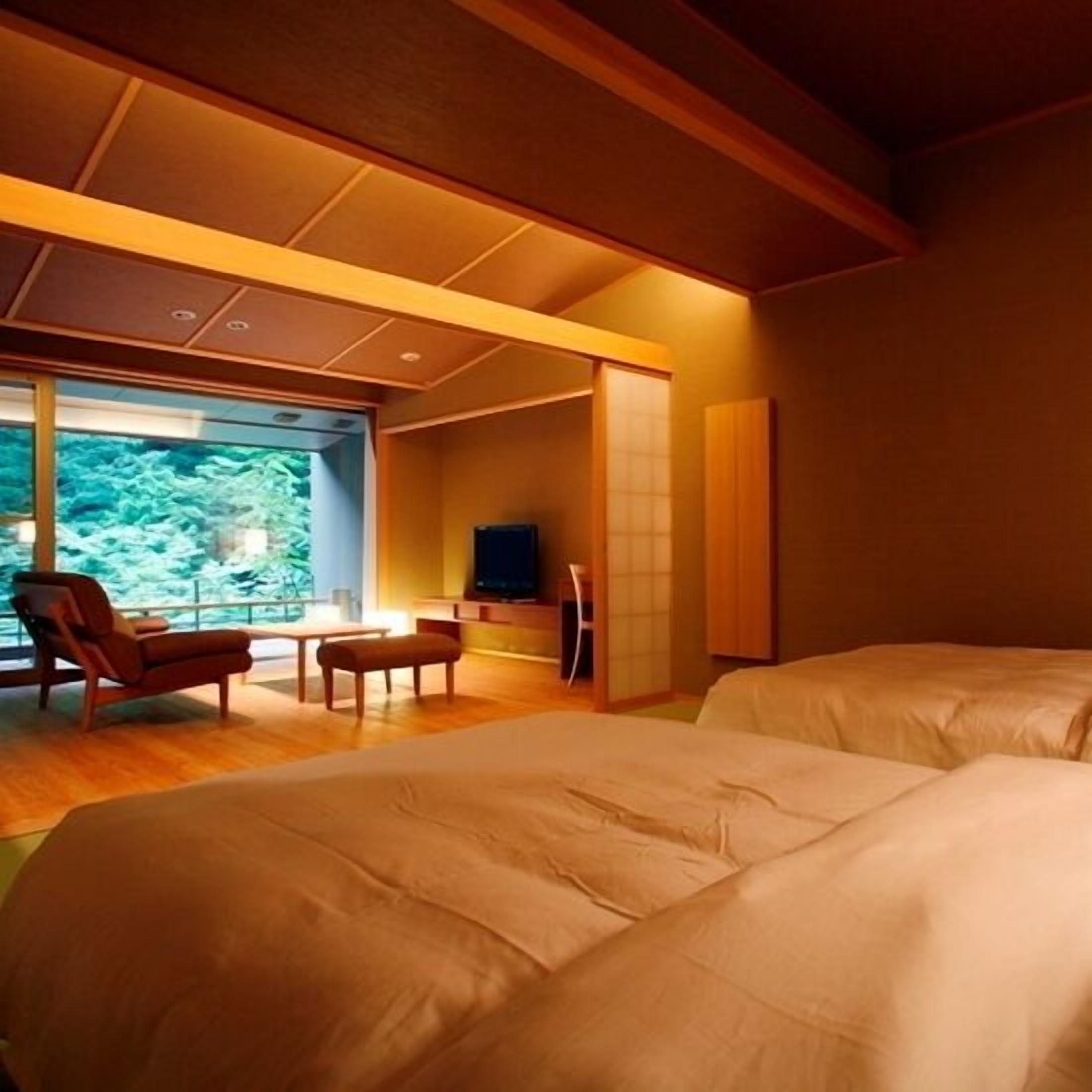 A room with two beds, a seating area with a chair and table, a television, and a large window with a view of trees.