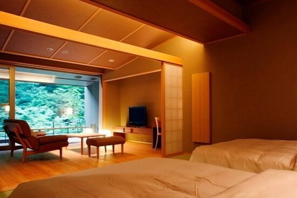 Une chambre avec deux lits, un coin salon comprenant une chaise et une table, une télévision et une grande fenêtre donnant sur la forêt.