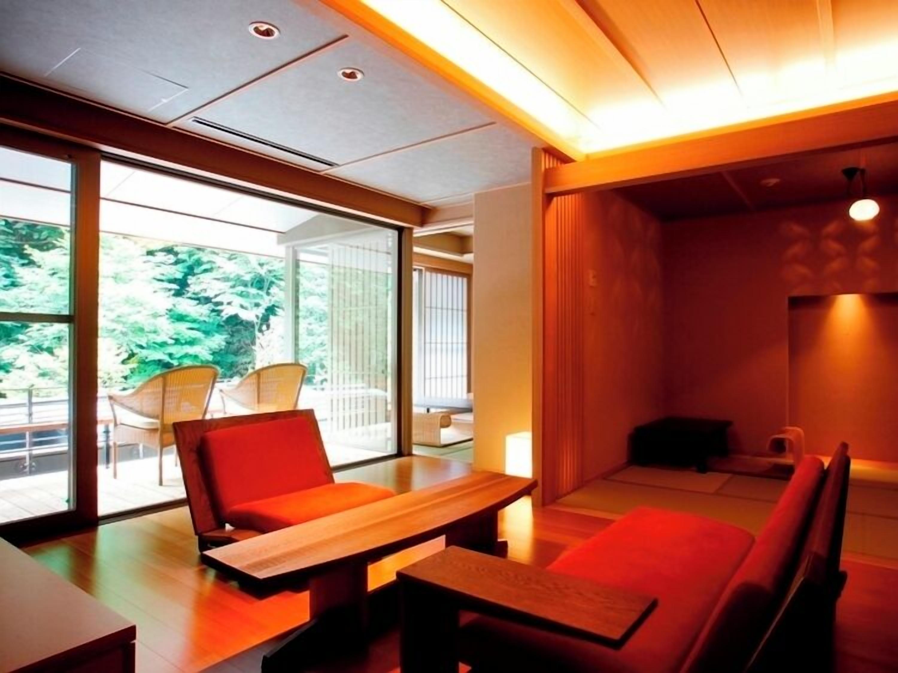 A modern interior with a dining area featuring a wooden table and red seating, large windows with a view of greenery, and a warm ambient light.
