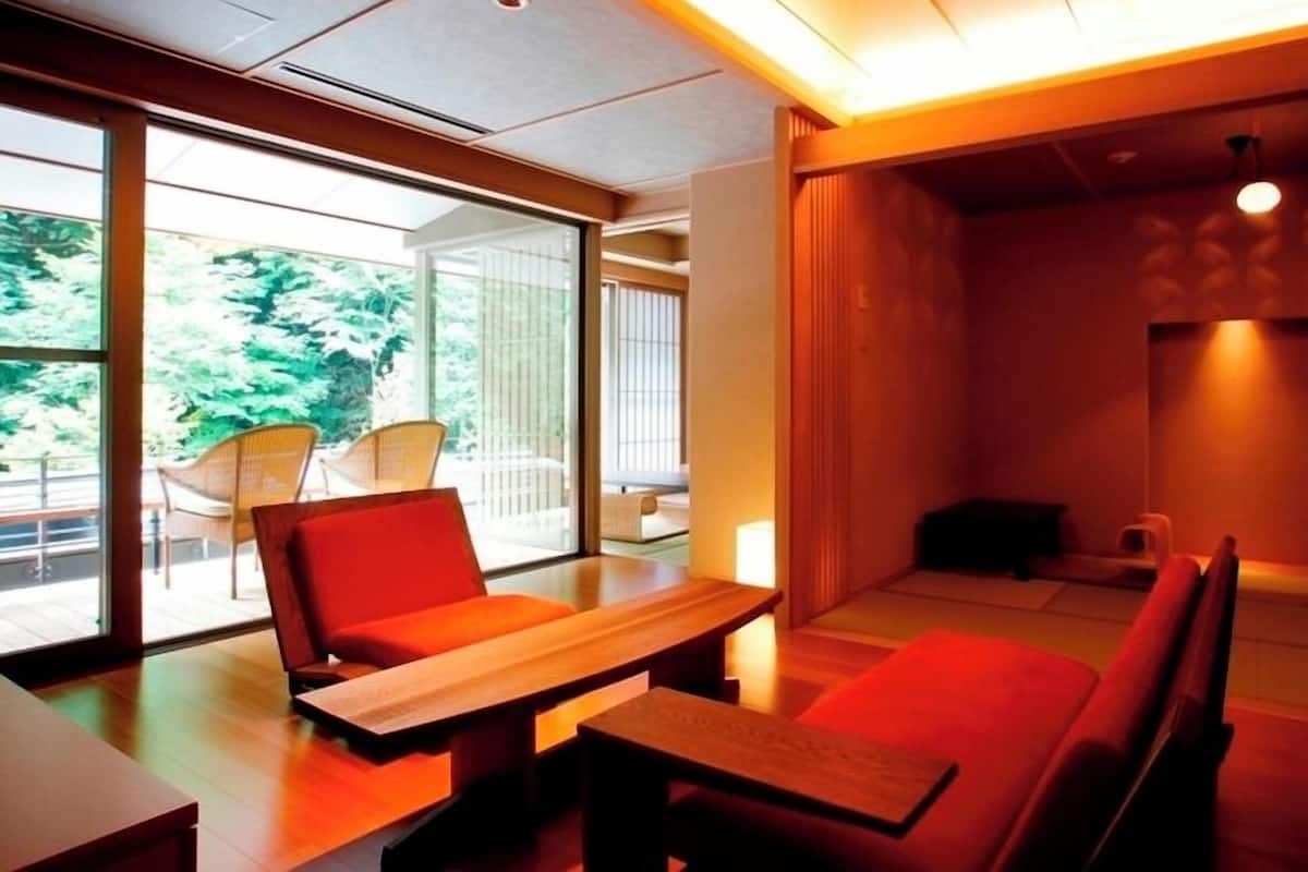 Un intérieur moderne avec un coin repas comprenant une table en bois et des sièges rouges, de grandes fenêtres offrant une vue sur la verdure et un éclairage d’ambiance chaleureux.