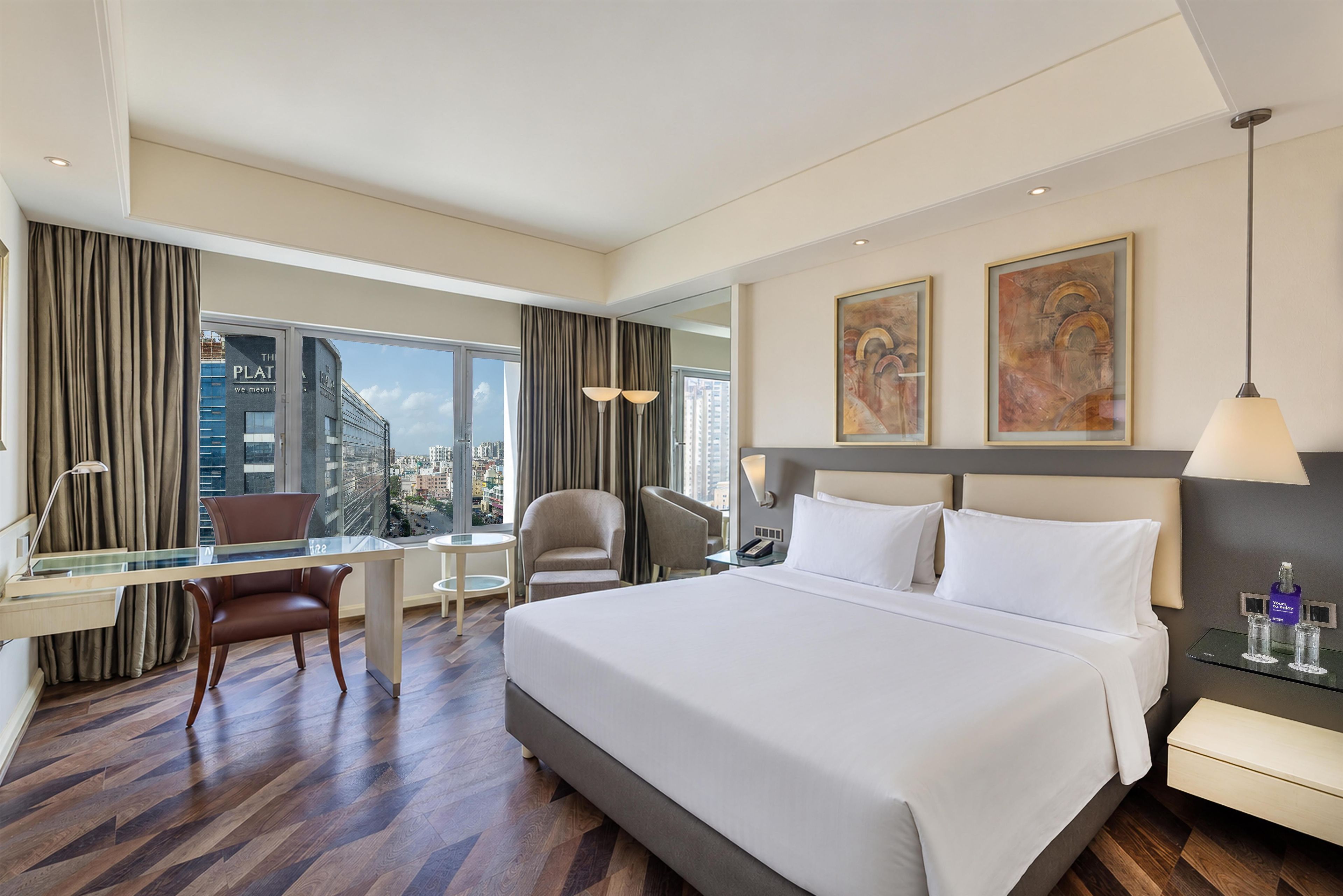 A hotel room with a large bed, a desk, a chair, a nightstand, and a view of the cityscape.