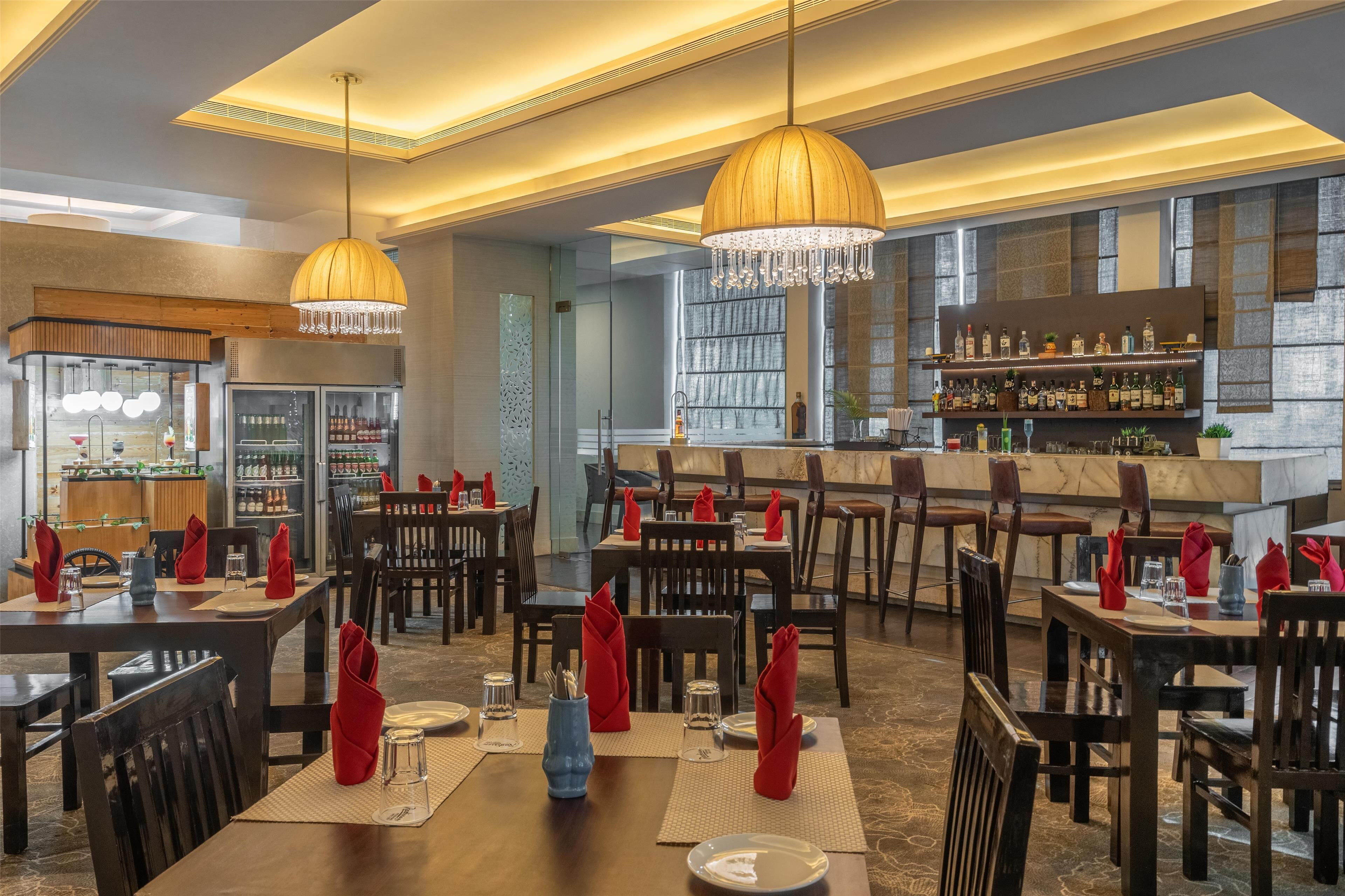 Een restaurant met meerdere tafels, rode servetten en een bar voorzien van flessen.