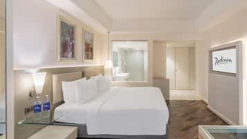 A hotel room with a large bed, bedside tables, a glass partition, a shower area, and a framed artwork on the wall.