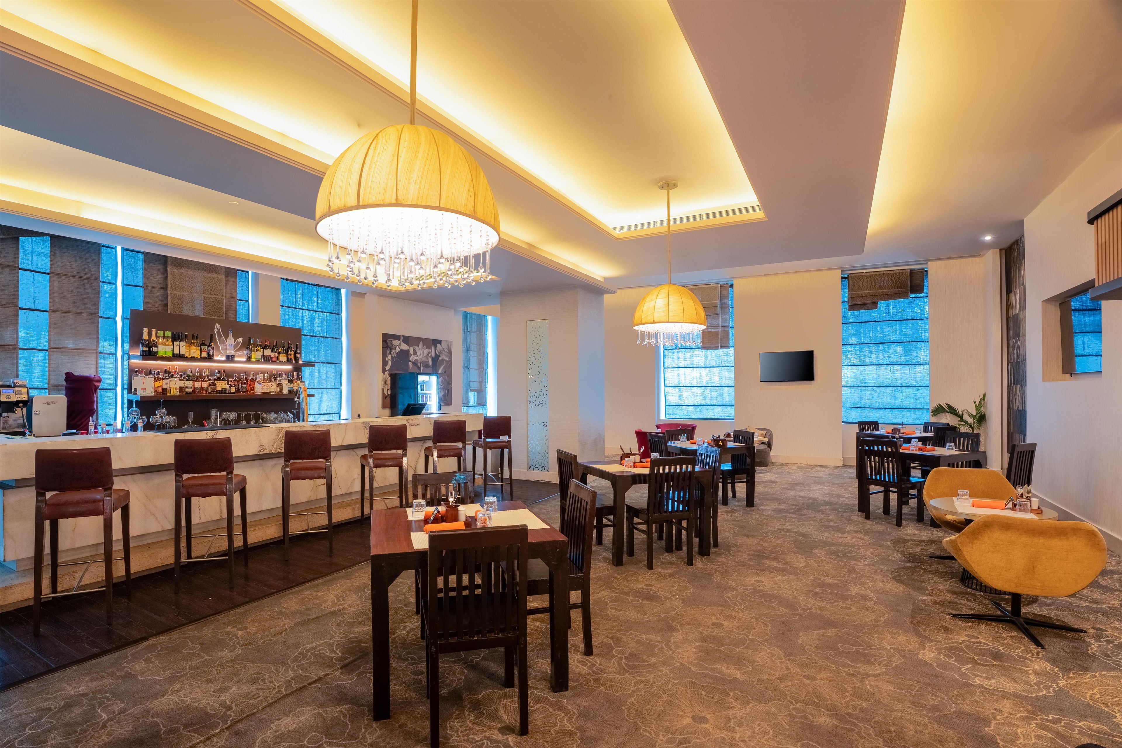 A modern bar area with a long counter, multiple bar stools, and a variety of bottles. There are dining tables with chairs arranged for guests.