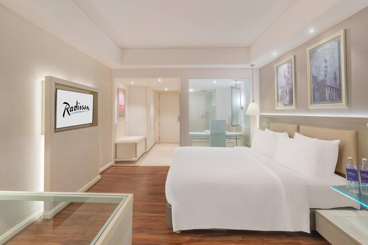 A hotel room with a large bed, a glass partition, a bathroom with a glass shower, and framed artwork on the walls.