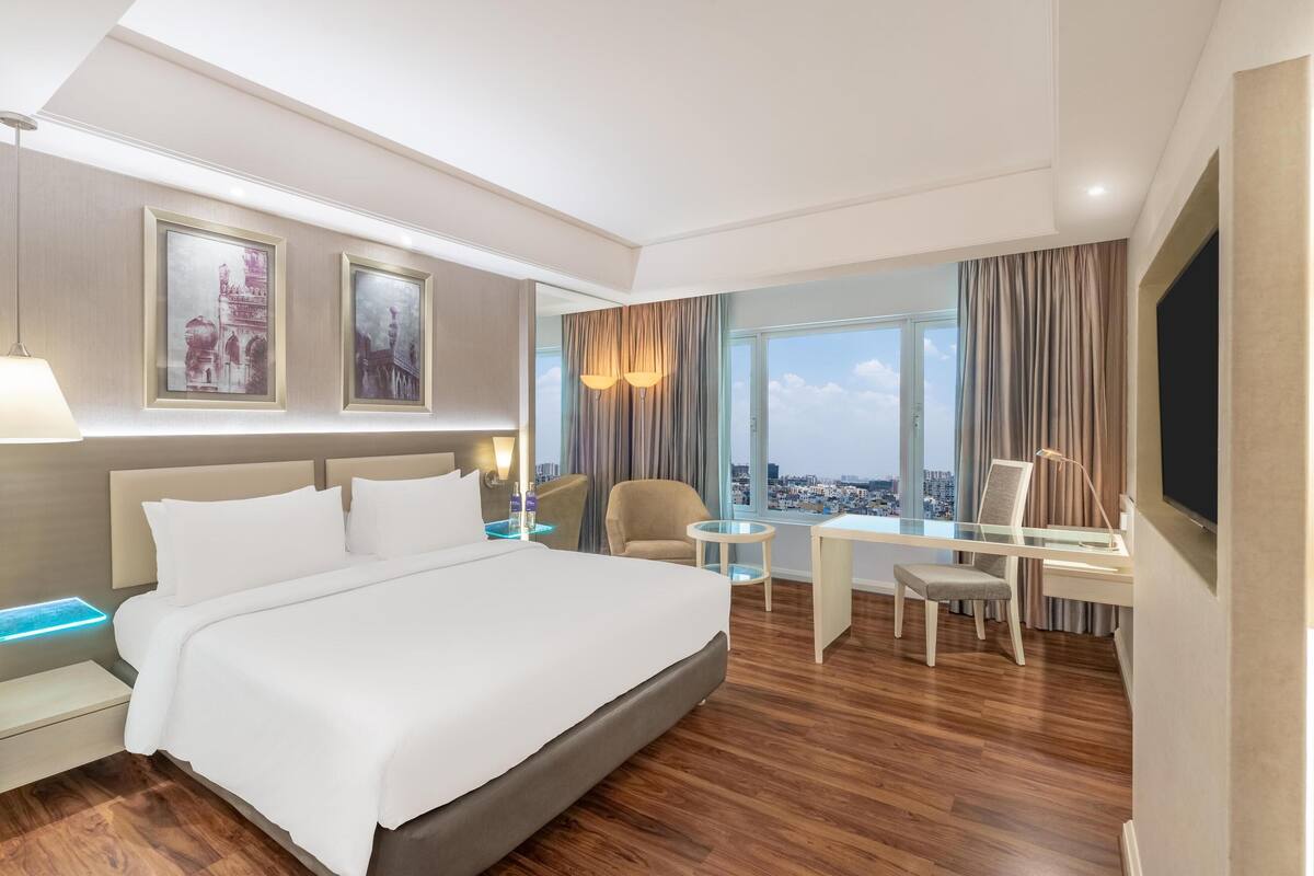A modern hotel room with a large bed, a desk, a chair, and a view of the city.
