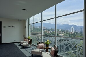 A spacious room with large windows, a sofa, a small table with a plant, and a view of a cityscape with mountains in the distance. - Hotel BH El Poblado (Medellín)