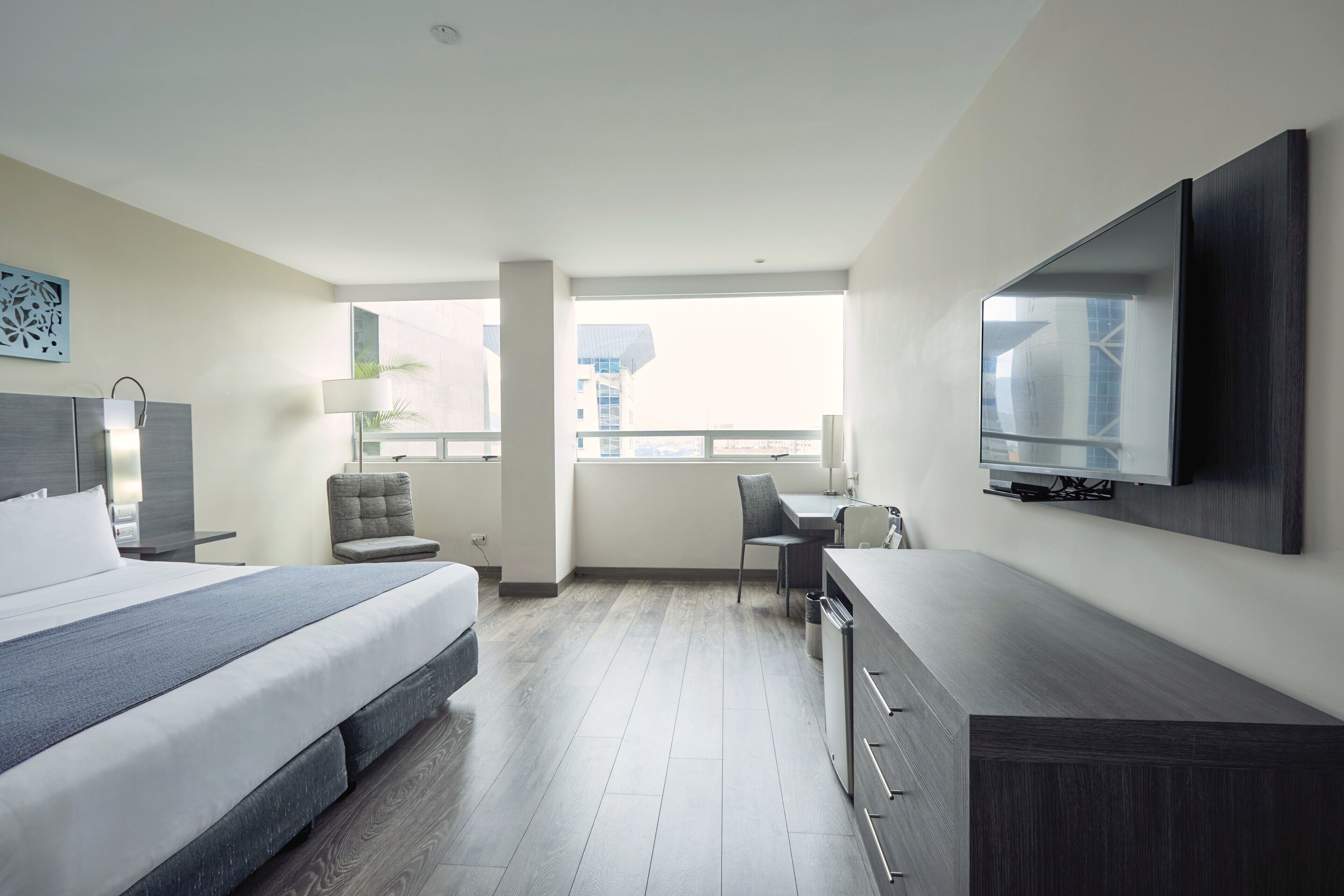 A modern hotel room with a bed, desk, chair, TV, and large windows.