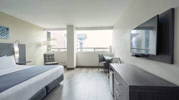 A modern hotel room with a bed, desk, chair, TV, and large windows.