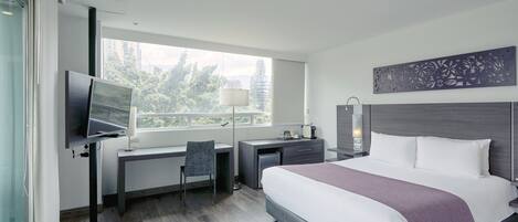 Une chambre d’hôtel moderne dotée d’un grand lit, d’un bureau, d’une chaise, d’une télévision et d’une fenêtre donnant sur des arbres et des immeubles.