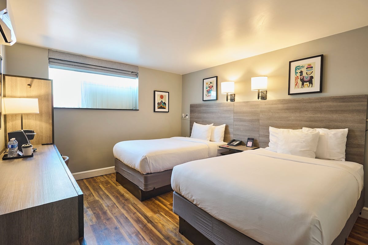A hotel room with two beds, a desk with a lamp, and artwork on the walls.