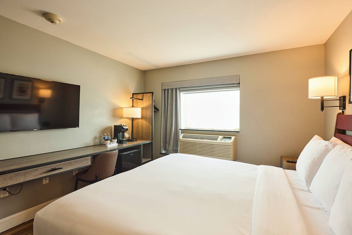 A hotel room with a large bed, a desk with a lamp, a flat-screen TV, and a window with curtains.