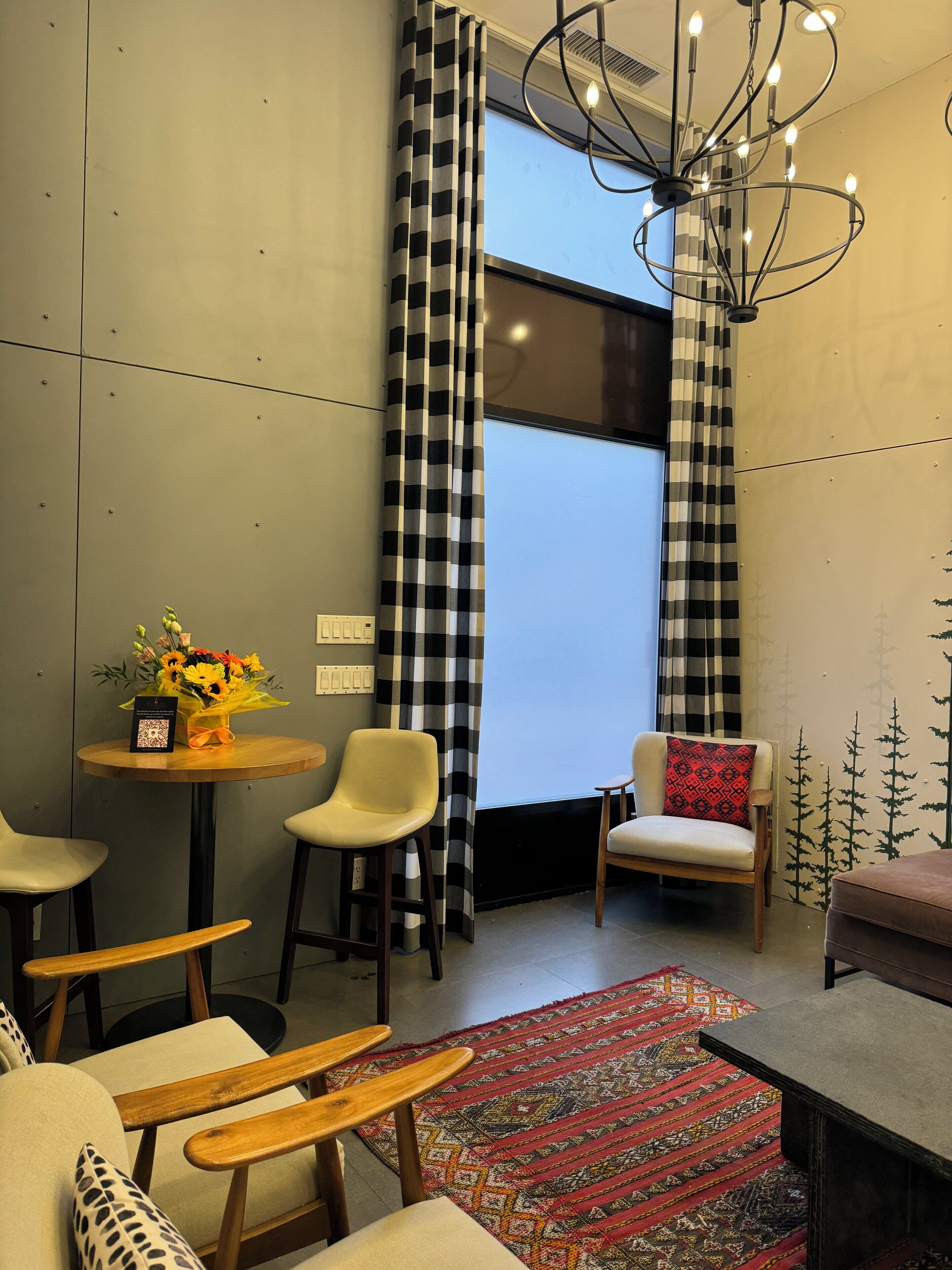 A modern room with a large window, checkered curtains, a chandelier, and a small table with a vase of flowers.