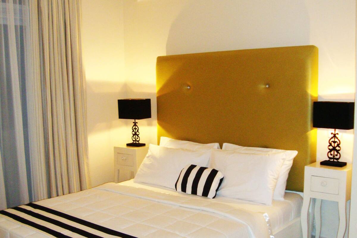A hotel room with a large bed, two bedside tables with lamps, a ceiling air conditioner, and a window with curtains.