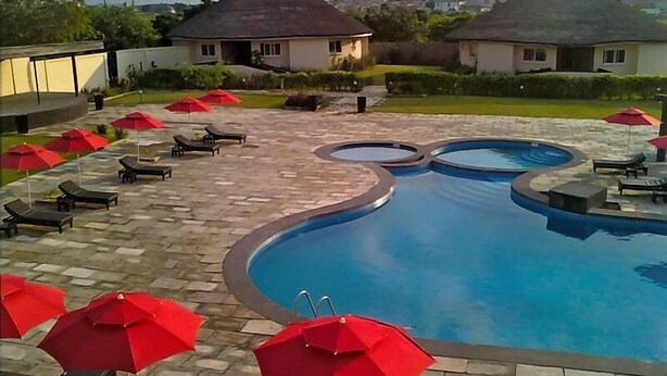 A swimming pool with lounge chairs and red umbrellas, surrounded by a paved patio area. - The Aknac Hotel (Accra)