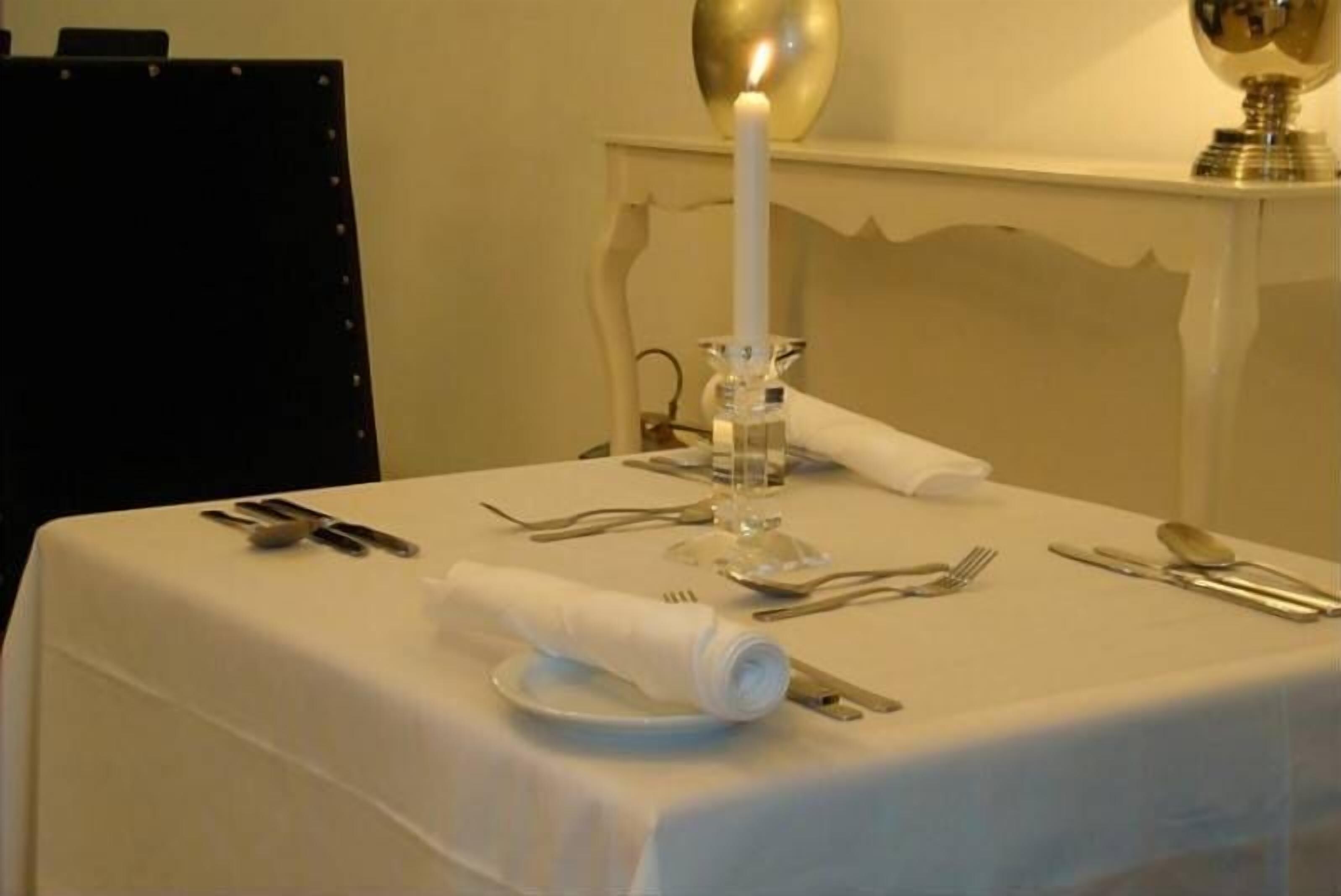Una mesa de comedor ordenada con mantel blanco, una vela encendida y cubiertos dispuestos para una comida.