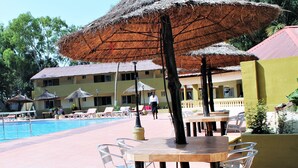 A pool area with lounge chairs and umbrellas, a dining table, and a building with a tiled roof in the background. - Badala Park Hotel (Serrekunda)