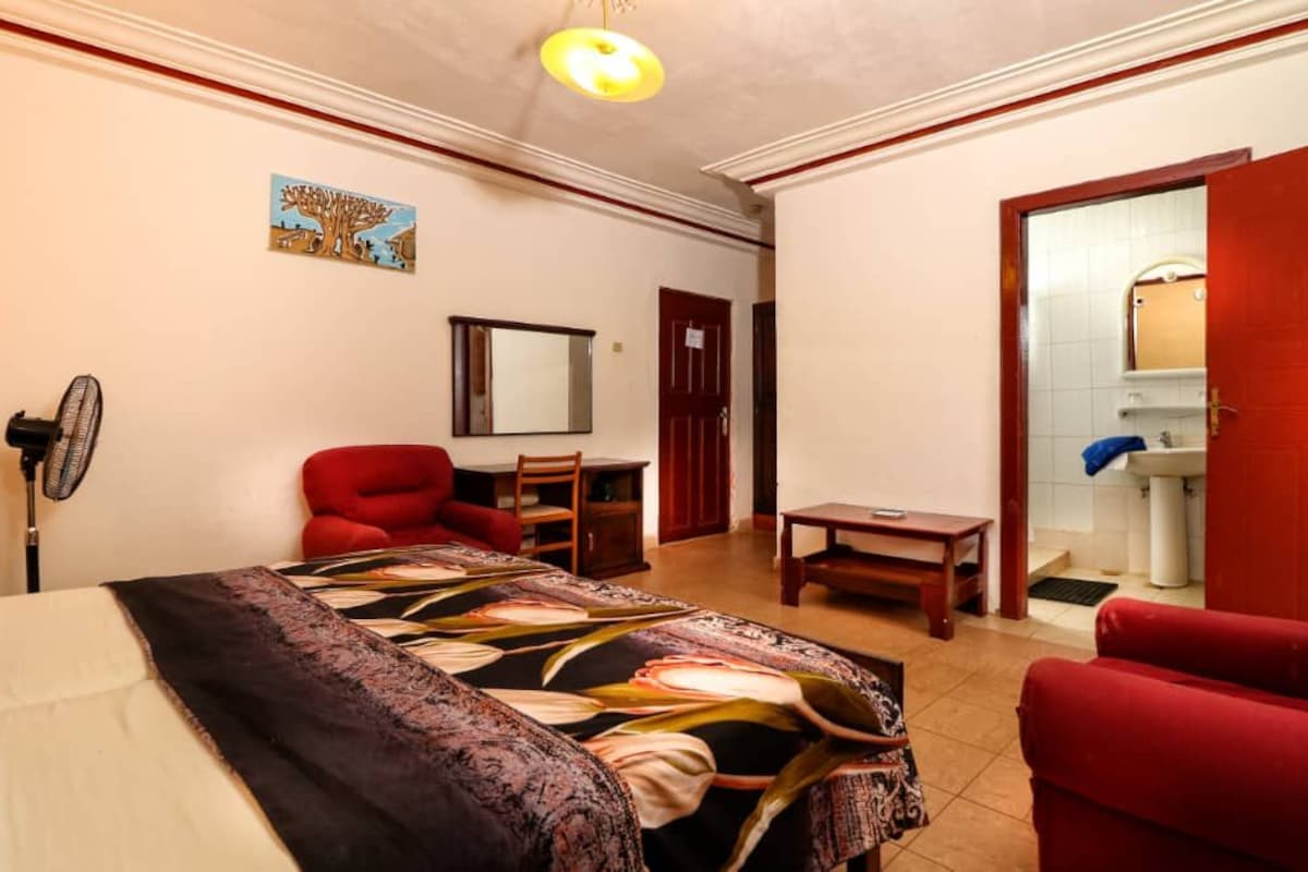 A hotel room with a bed, a desk, a chair, a red sofa, a fan, a mirror, and a bathroom visible through an open door.