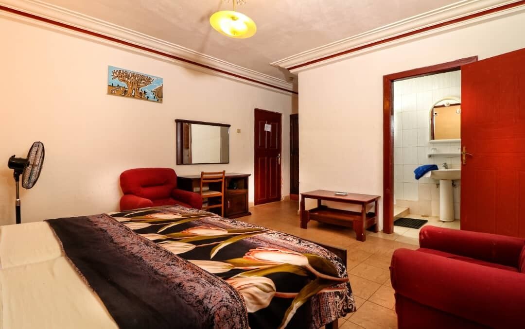 Een kamer met een bed, een rode stoel, een klein tafeltje, een ventilator en een deur naar een badkamer.