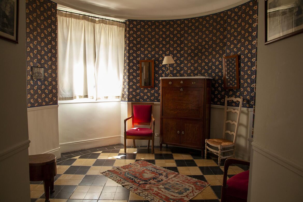 A room with patterned wallpaper, a checkered floor, a red chair, a wooden cabinet, and a window with curtains.