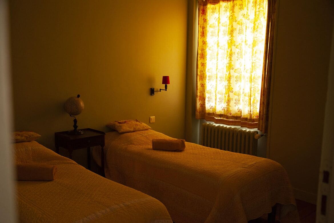 A room with two beds, a lamp, a wall-mounted light, and a window with curtains.