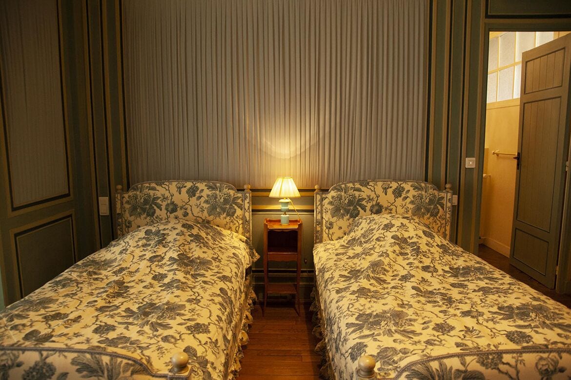 A hotel room with two beds, a nightstand with a lamp, and a door to the right.