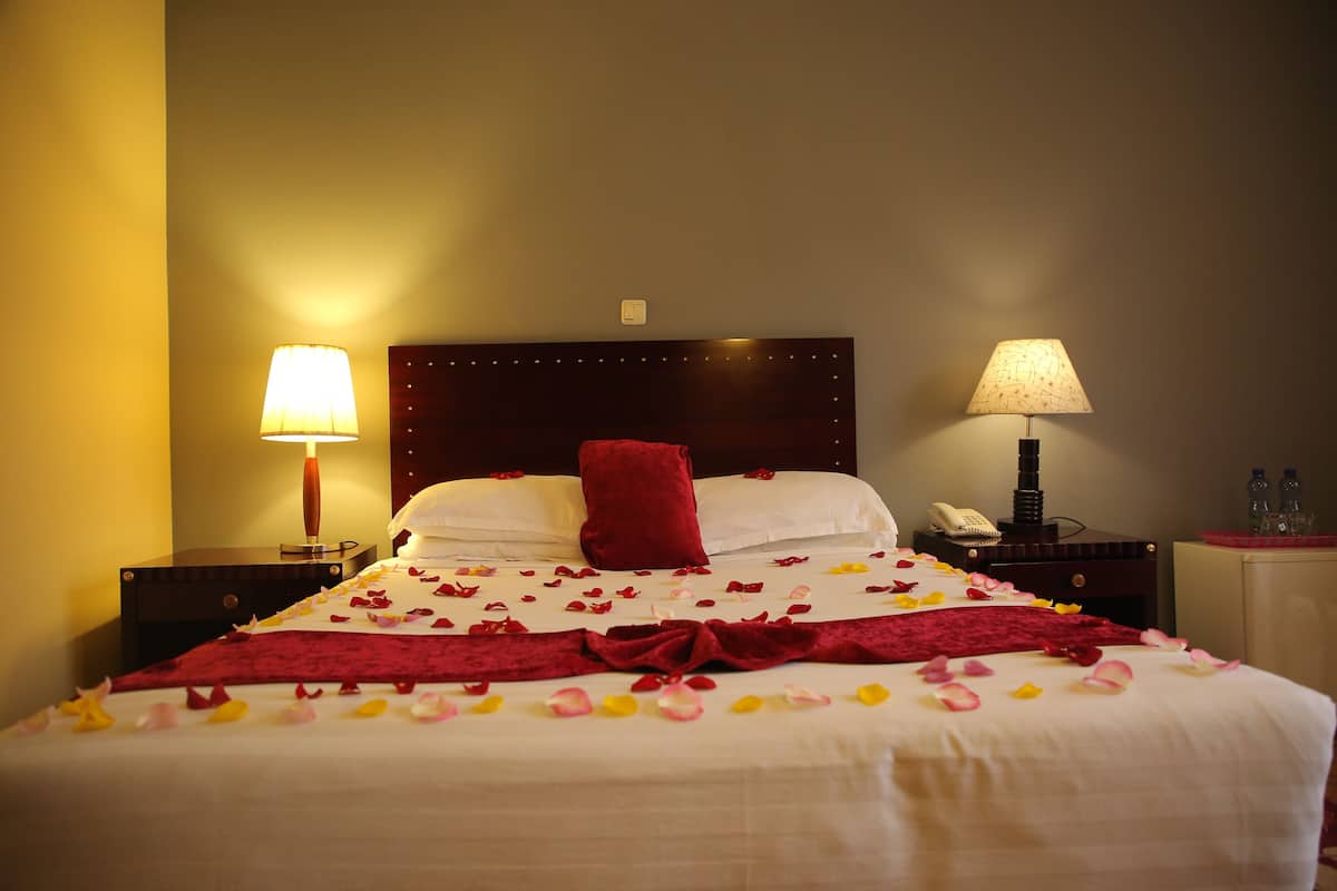 Una cama bien hecha con un almohadón rojo, sábanas blancas y una manta roja. Hay dos mesitas de noche con lámparas, un teléfono y un pequeño mueble con botellas.