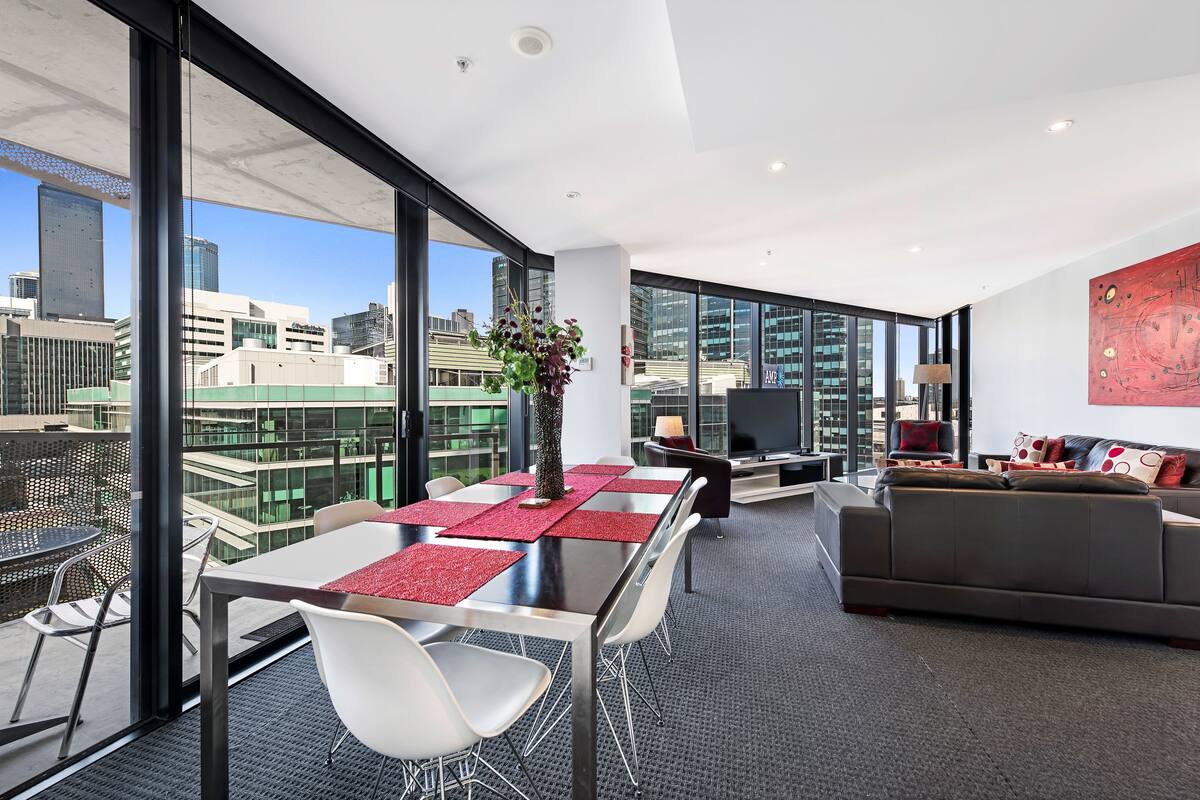 A modern hotel room with a dining area, a sofa, and a large window offering a city view.