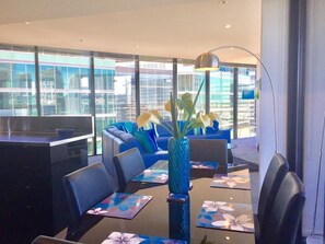 A modern dining area with a glass table, blue upholstered seating, and a vase with flowers. - Docklands Executive Apartments (Docklands)