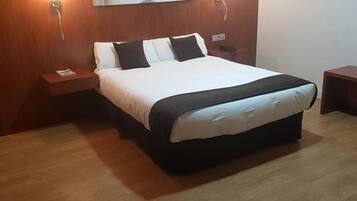 Una habitación de hotel ordenada con una cama grande, dos mesitas de noche, una lámpara de pared y una pequeña mesita de noche con lámpara.