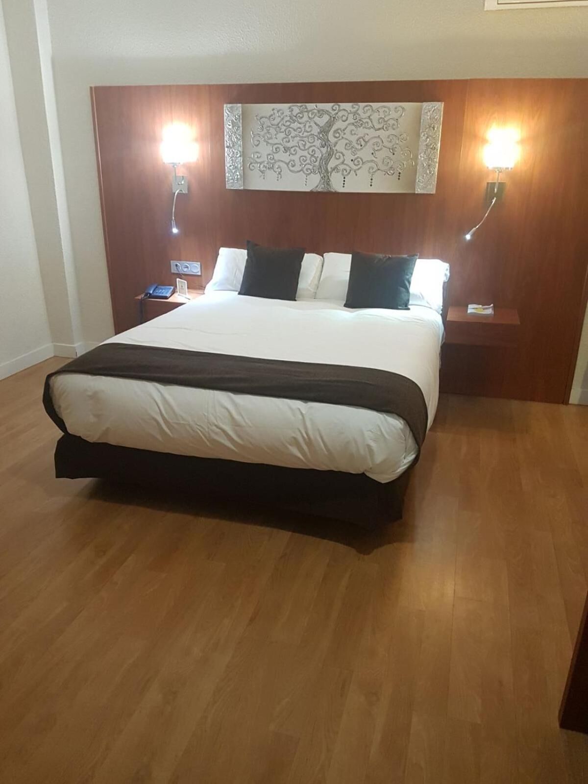 Habitación de hotel con una cama grande, suelo de madera, una mesita de noche con teléfono y lámparas de pared.