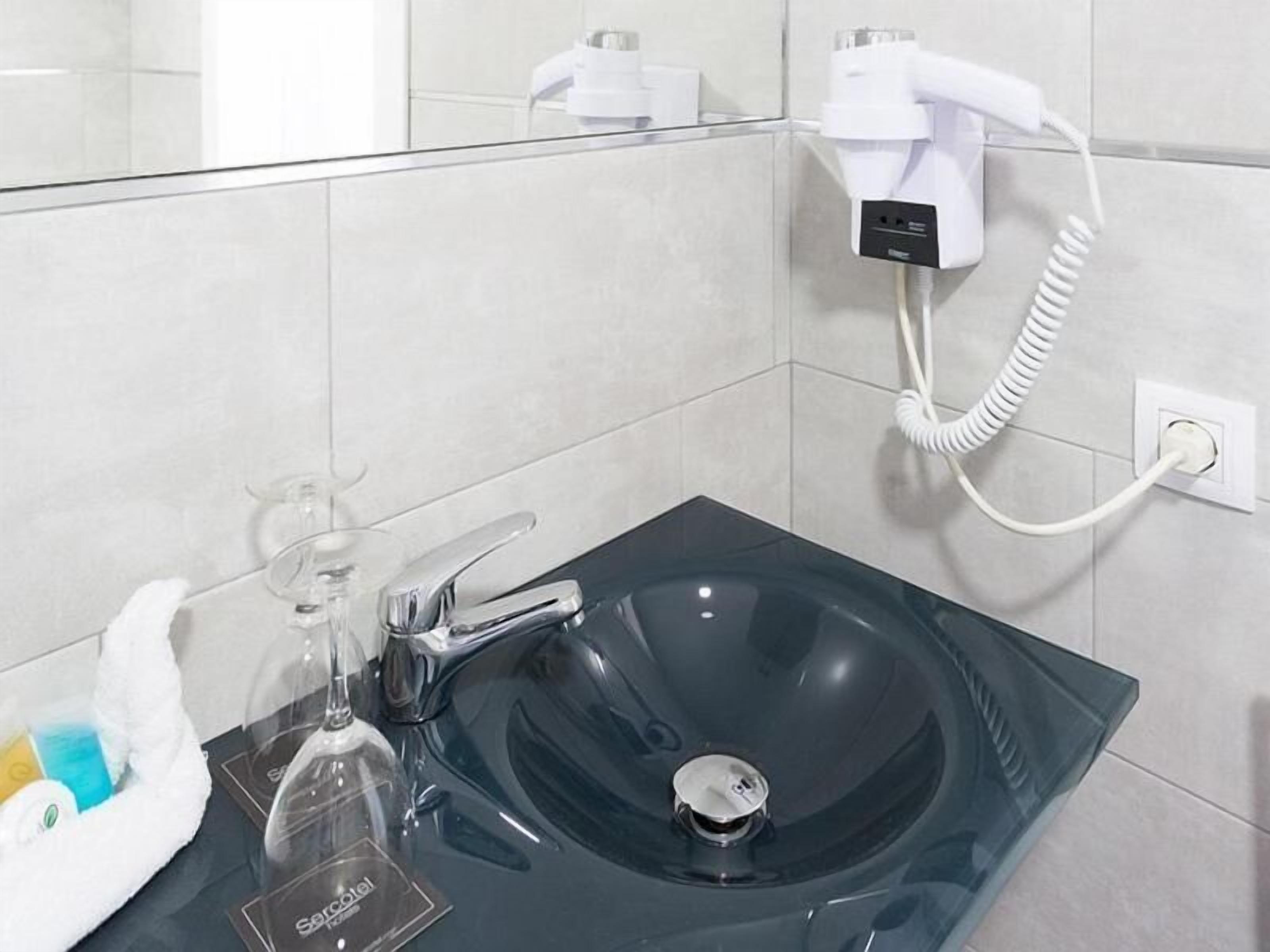 Un baño moderno con un lavamanos de vidrio, un cabezal de ducha manual blanco y una botella de jabón de manos.