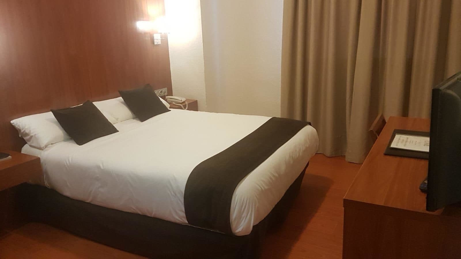 A hotel room with a large bed, a bedside table, a lamp, and a television.