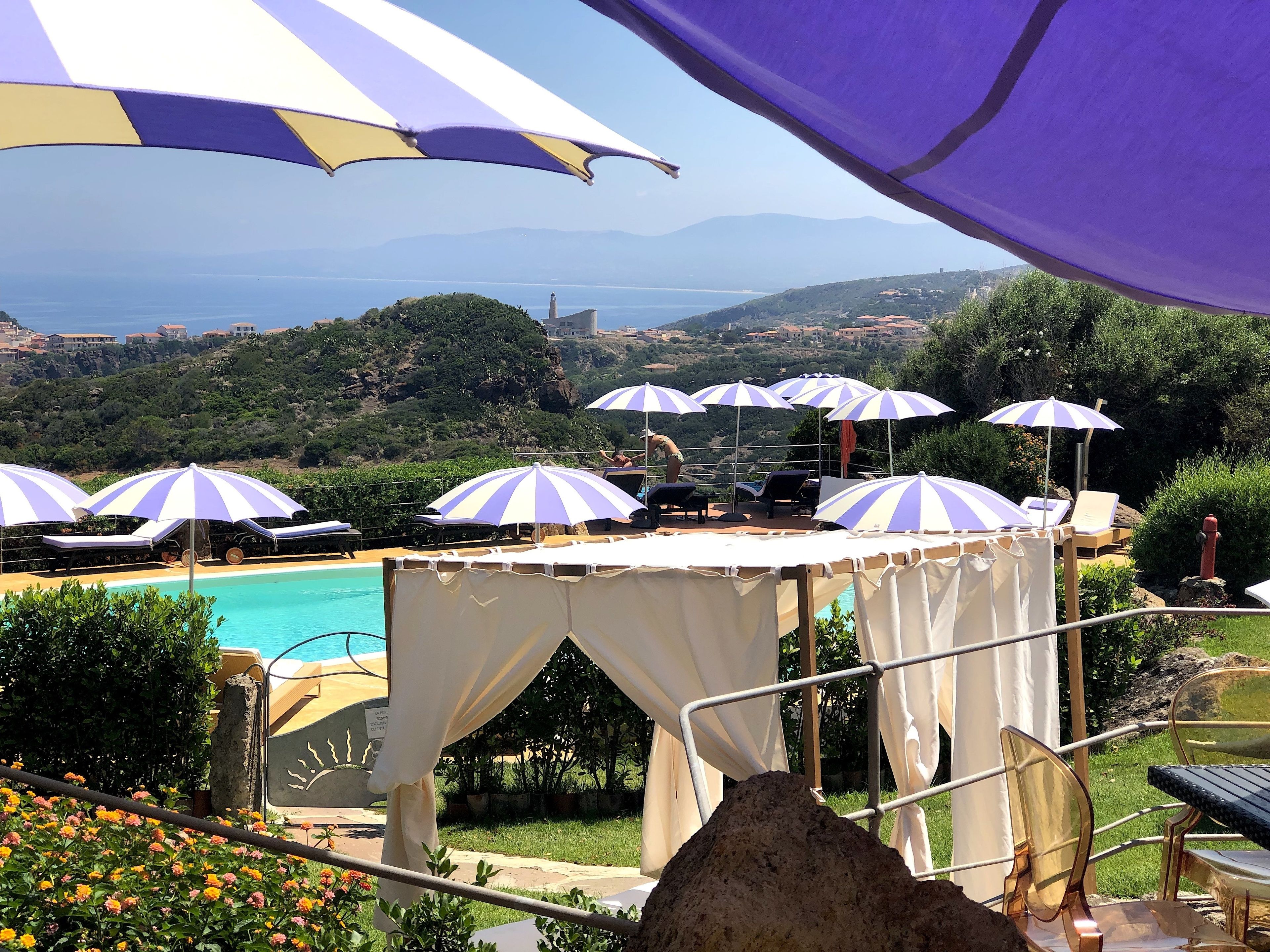 A pool area with multiple sun umbrellas and lounge chairs, surrounded by greenery and a view of the sea.