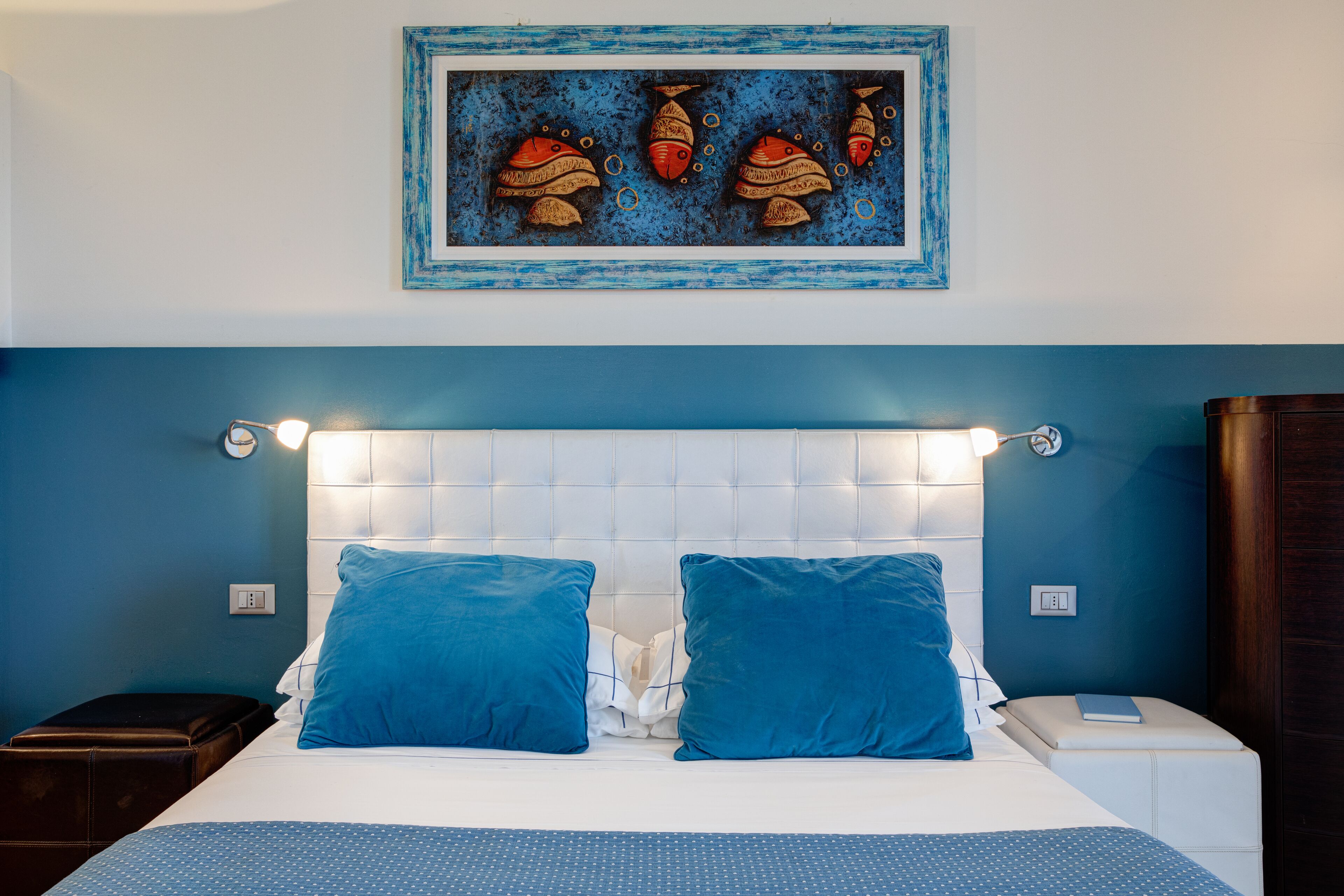 A bedroom with a blue headboard, two blue pillows, a white bedspread, and a wall art of fish.