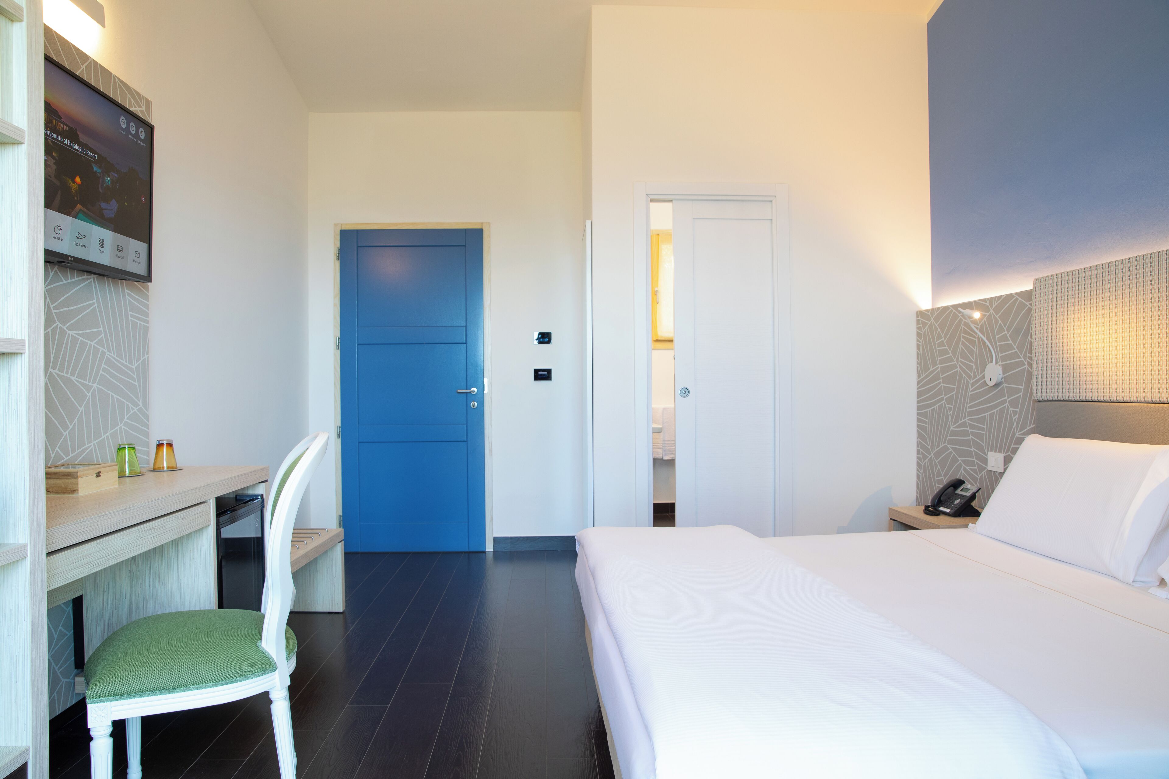 A hotel room with a bed, a desk with a chair, a blue door, and a wall-mounted TV.